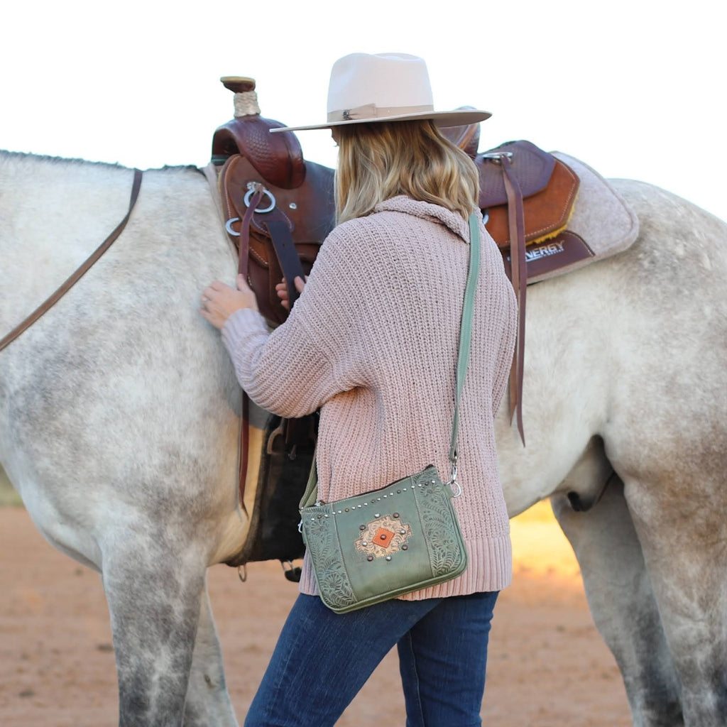American West Handcrafted Leather crossbody bag with floral tooled leather details and western styling worn outdoors beside horse