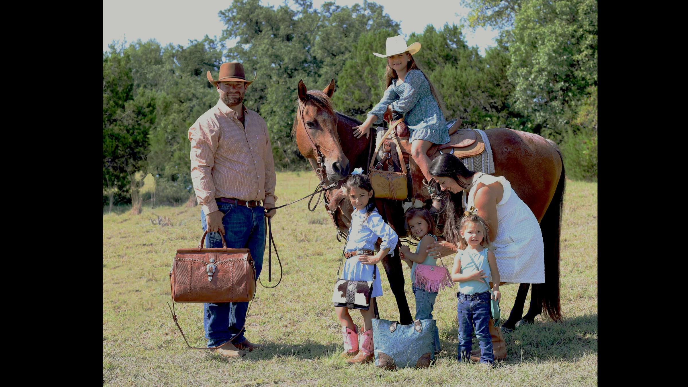 Family outdoors with horse showcasing American West Handcrafted Leather western handbags, floral tooled leather duffle bag, crossbody purses, and tote bags in a ranch setting