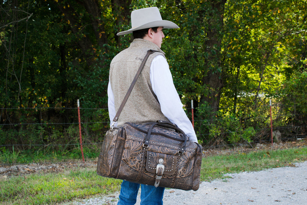 Lifestyle with model of Classic Travel duffel bag with front pouch and side zipper pockets in distressed charcoal brown with hand tooled leather and buckle set